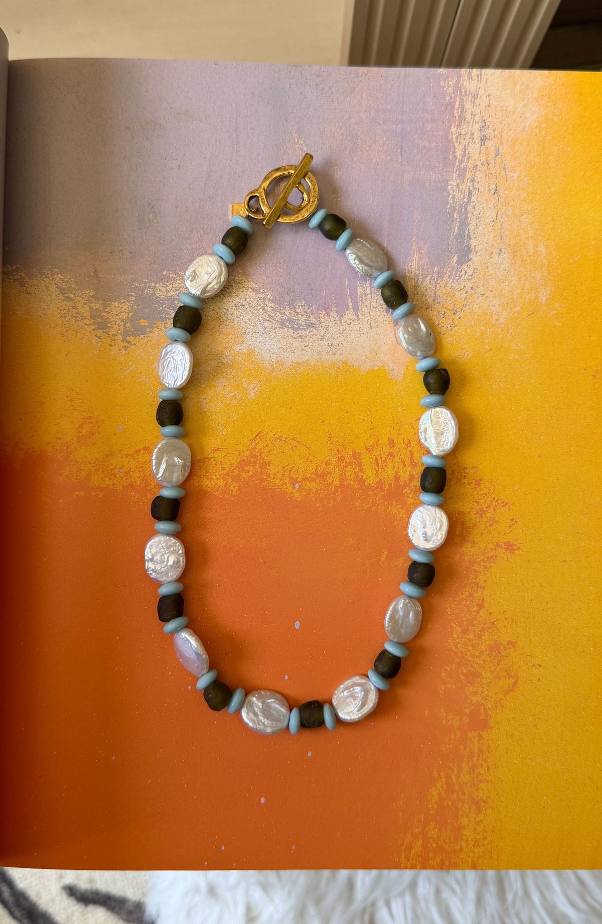 A beaded necklace with flat white pearls, dark brown beads, light blue spacers, and a gold toggle clasp lies on an orange and yellow painted background.