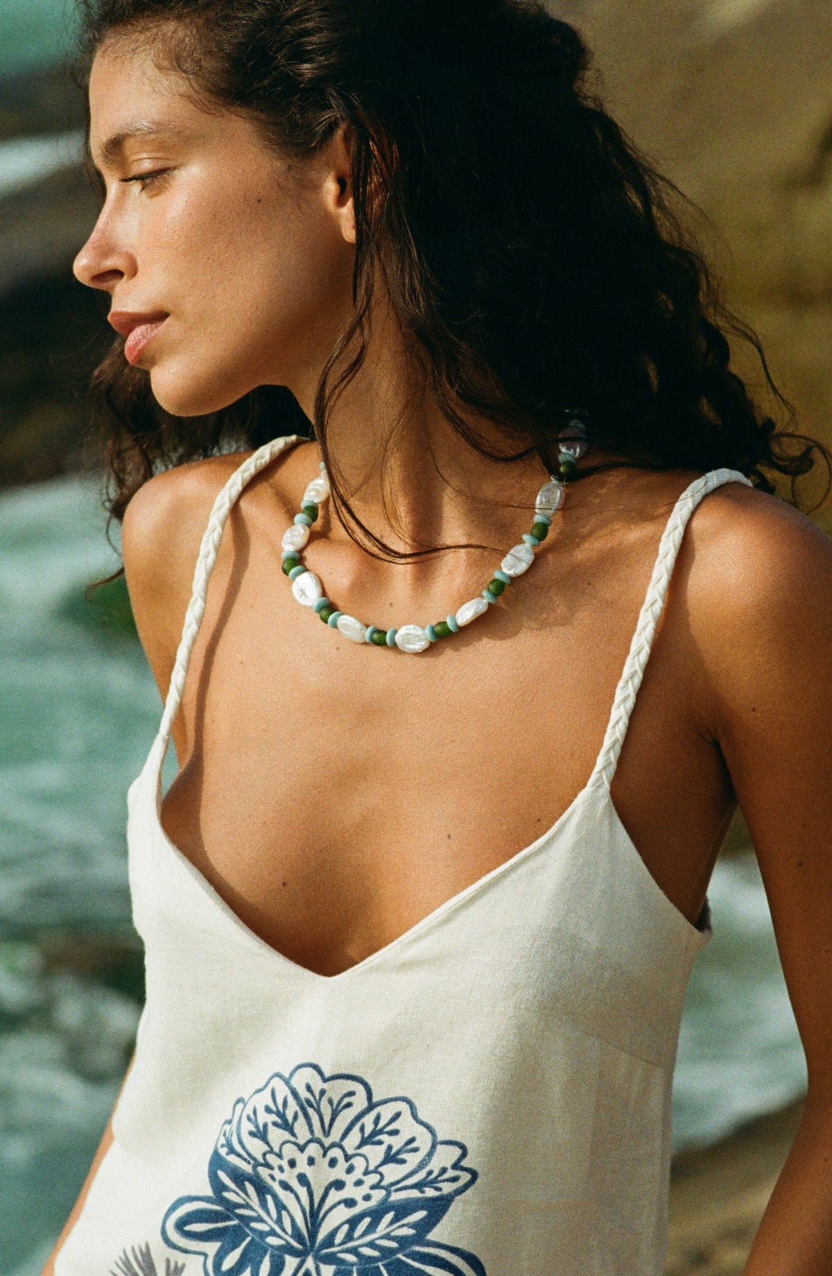 A woman wears a beaded necklace of white, green and turquoise beads with a cream top featuring a blue floral print.