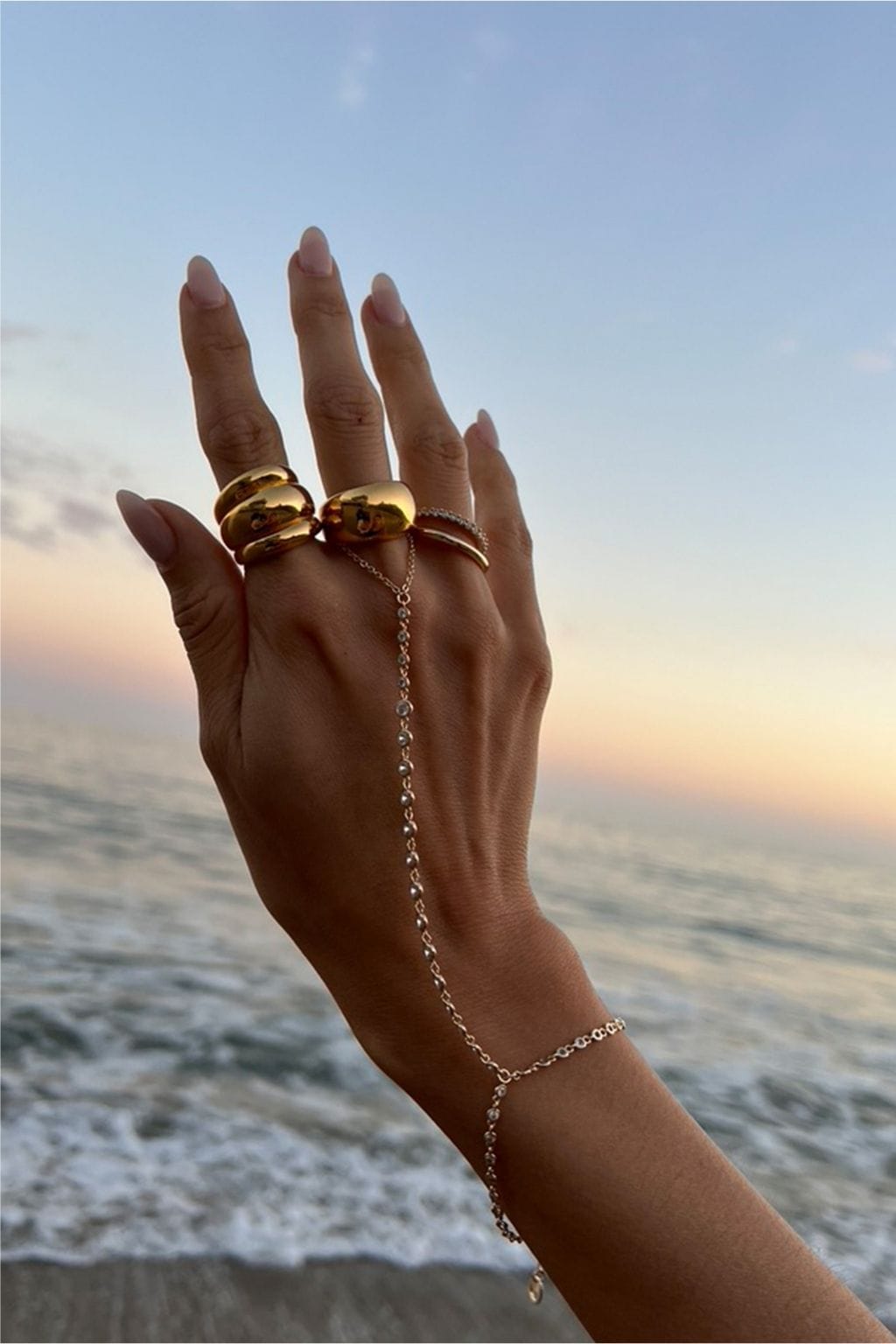A raised hand wears a delicate gold hand chain with small crystals, plus chunky gold rings, against an ocean sunset.