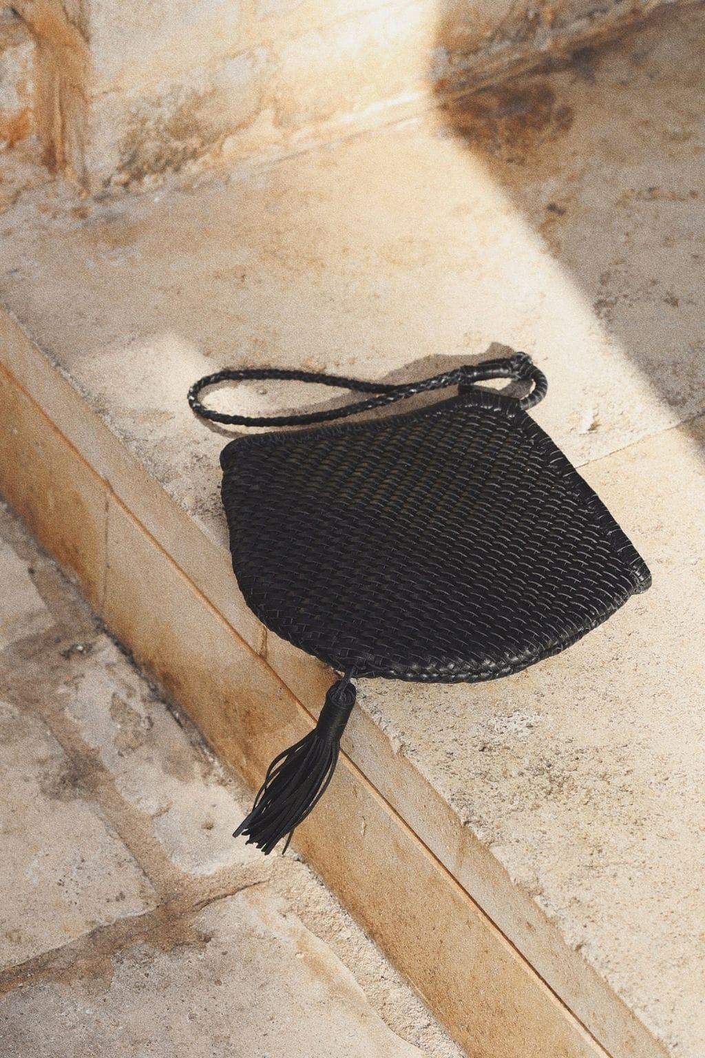 Black woven clutch bag with braided handle and tassel charm, displayed on stone surface.