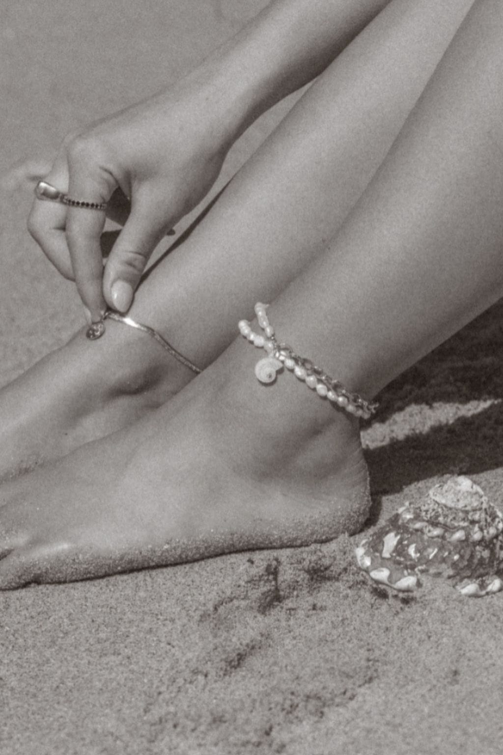 Pearl beaded anklet with charm detail worn on ankle, shown in black and white beach setting.