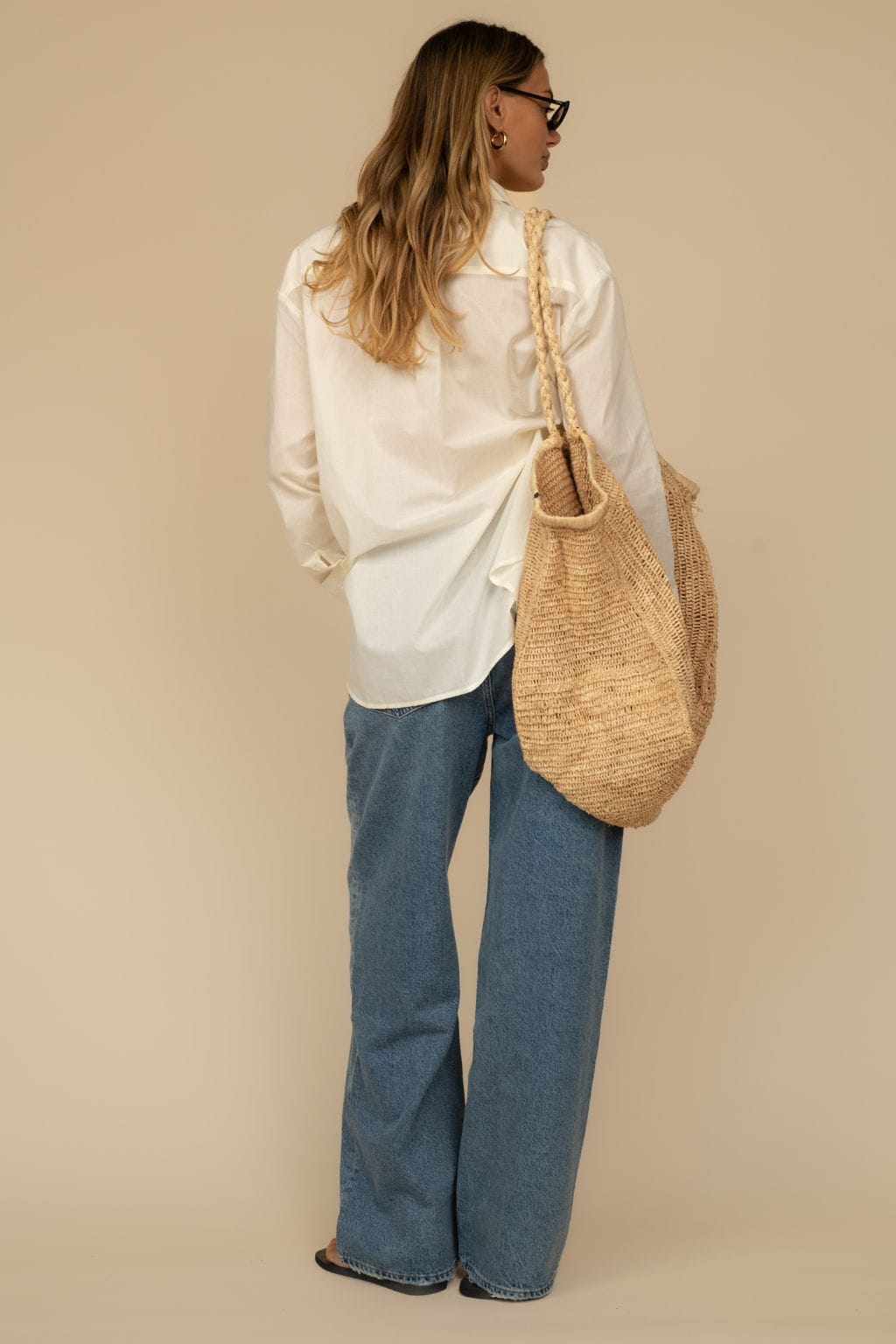 Woman wearing cream pleated baggy pants with white button-up shirt and tan woven shoulder bag.
