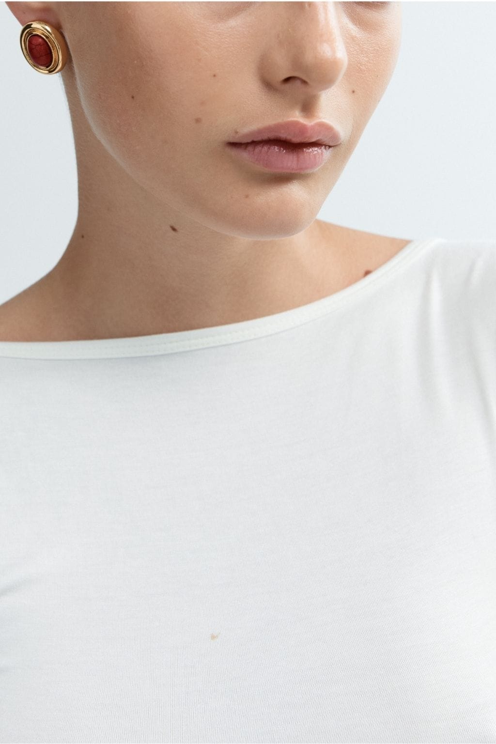 Person wearing burgundy and gold oval stud earring on ear while wearing white crewneck shirt.