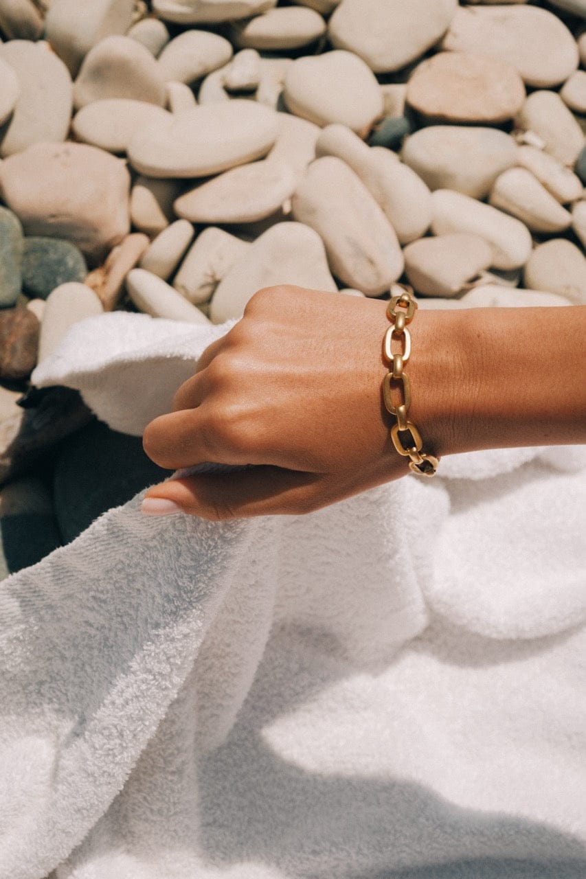 Gold chain link bracelet worn on wrist over white fabric with pebbles in background.