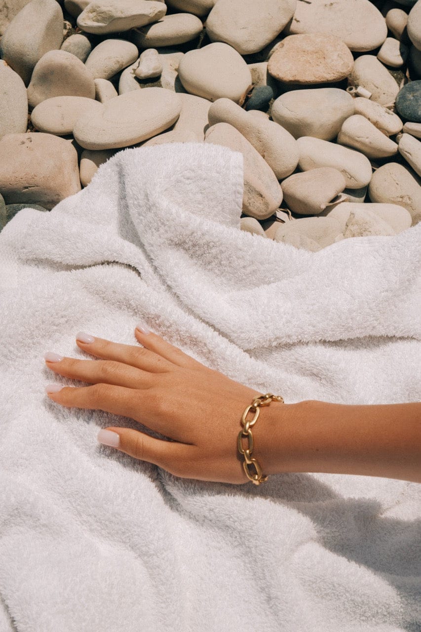 Gold chain link bracelet worn on wrist against white cloth and pebble background.