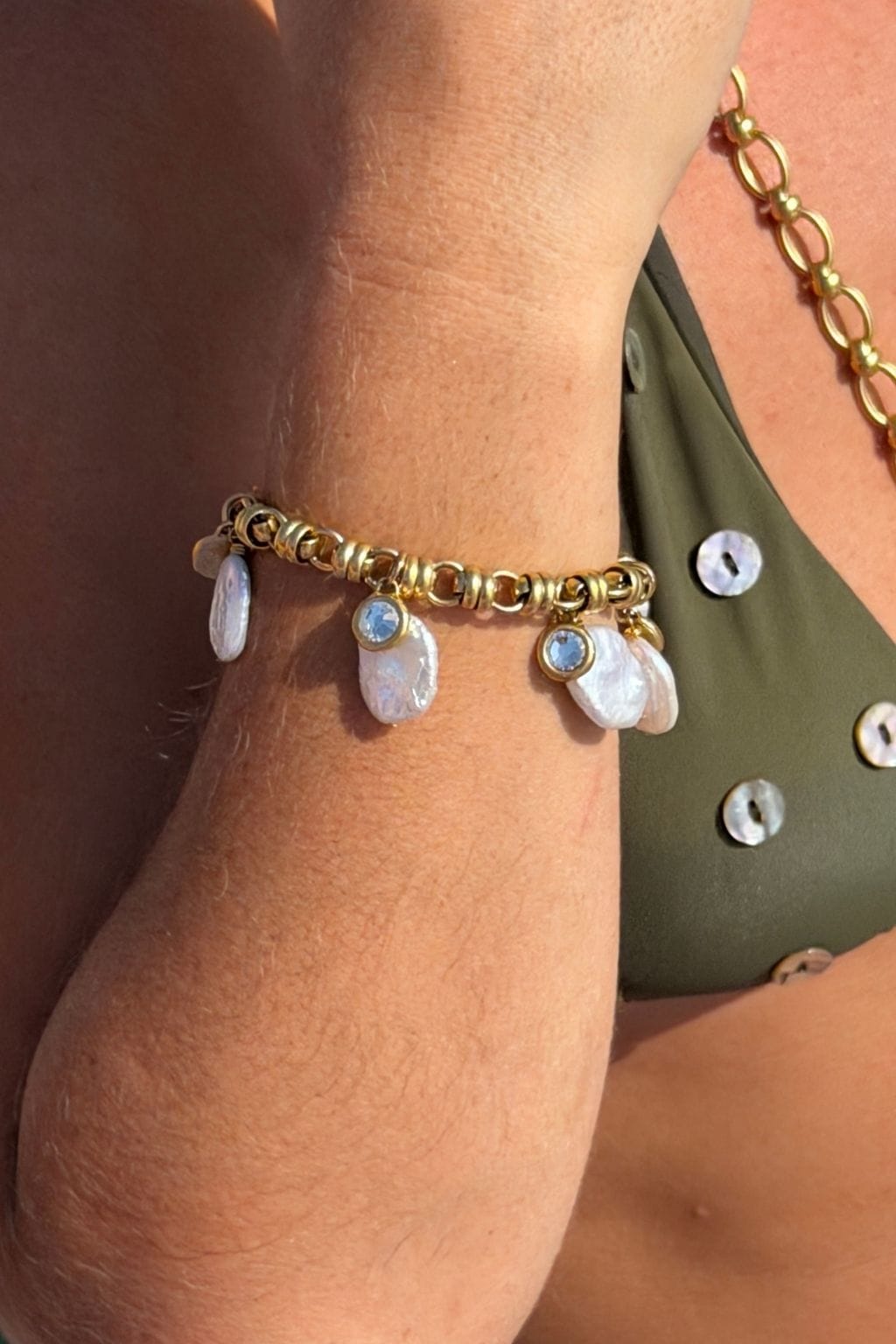 A gold chain bracelet worn on a wrist, with flat white pearl charms and small round light blue stones hanging from it.