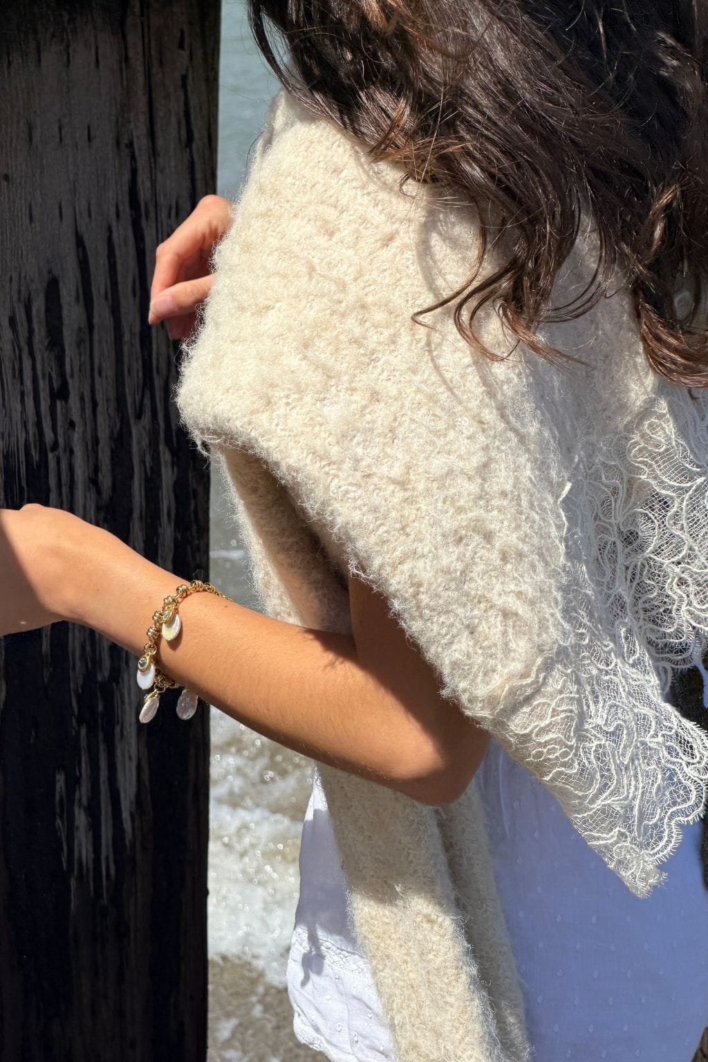 A woman wearing a gold bracelet with small pearl-like charms rests her arm near a dark wooden post, a cream knit sweater draped over her shoulders.