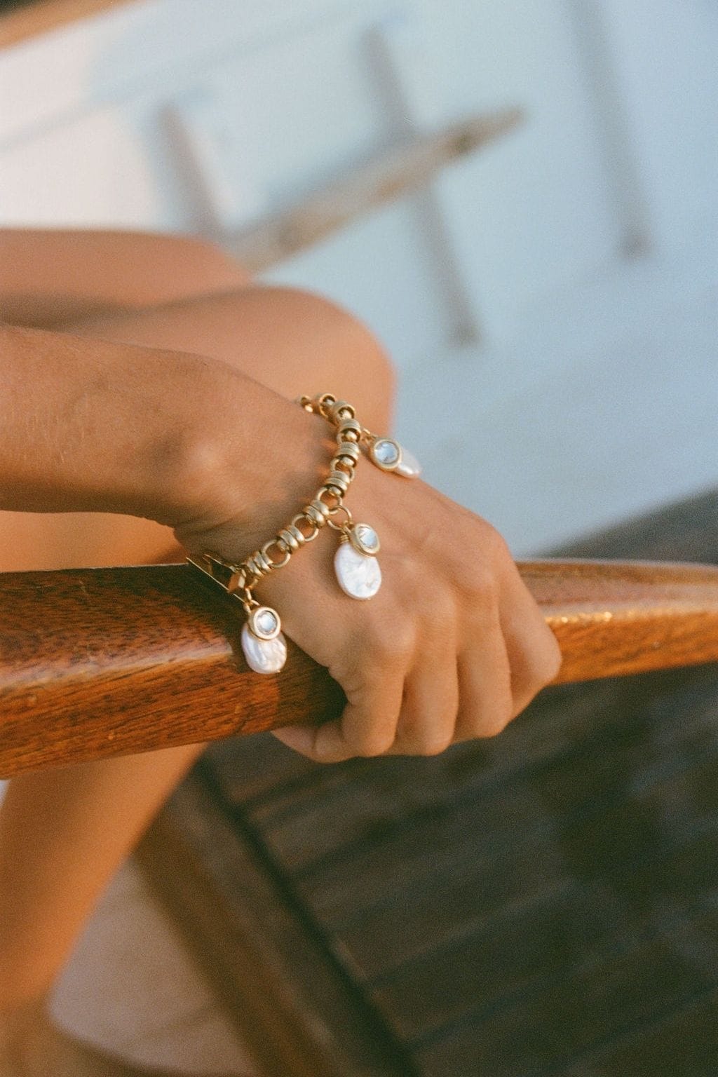 A gold chain-link bracelet with dangling flat white pearl charms is worn on a wrist resting on a wooden railing.