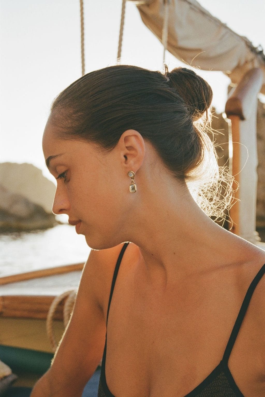 A woman in profile with her hair in a bun wears a small gold drop earring with clear stones on a boat.