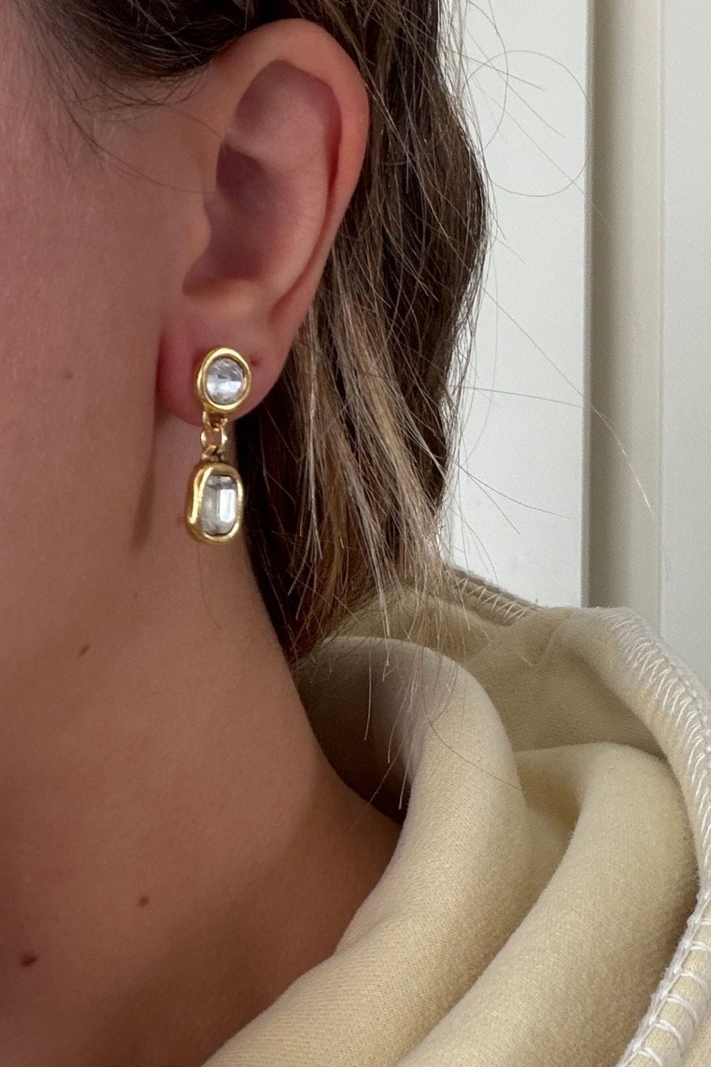 A gold drop earring with two clear crystal stones, one round and one rectangular, worn on an ear beside a cream collar.