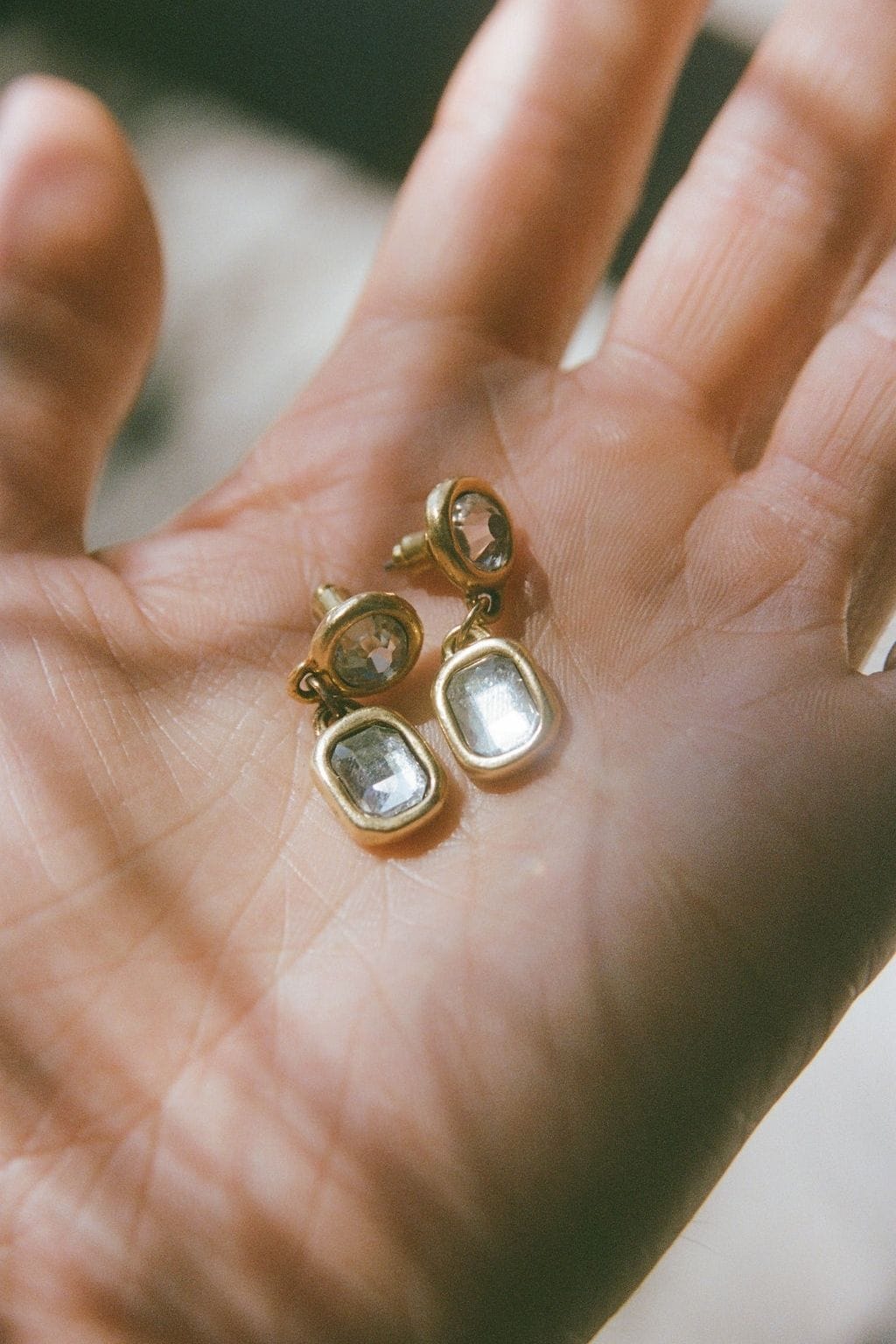 An open palm holds a pair of gold-toned drop earrings, each with a round and a rectangular clear crystal.