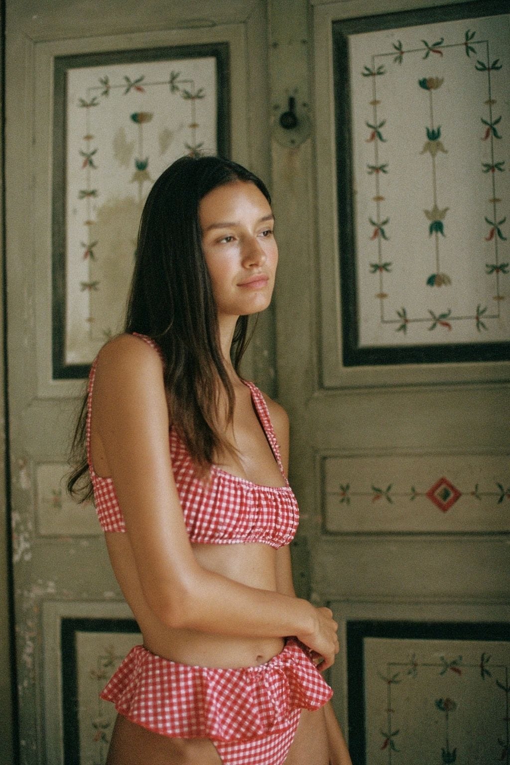 Red and white gingham bikini top with tie-shoulder straps on model against patterned wall background.