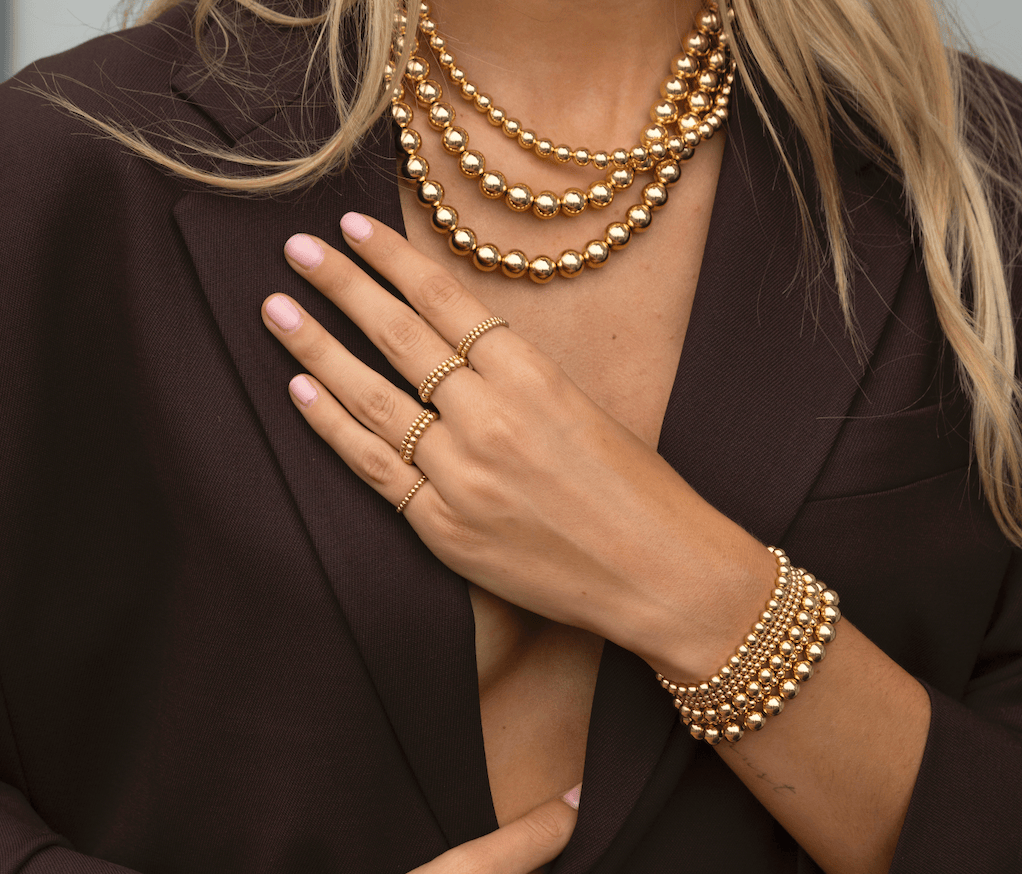 Gold beaded bracelet worn on wrist with matching beaded necklaces and rings.