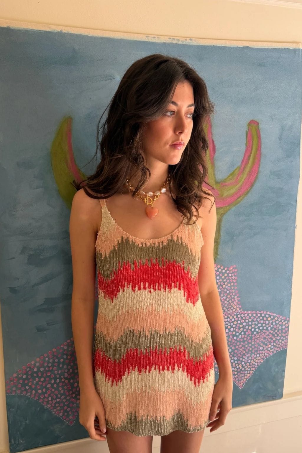 A woman with wavy dark hair wears a gold chain necklace with pearls and a peach heart pendant, standing before a painting.