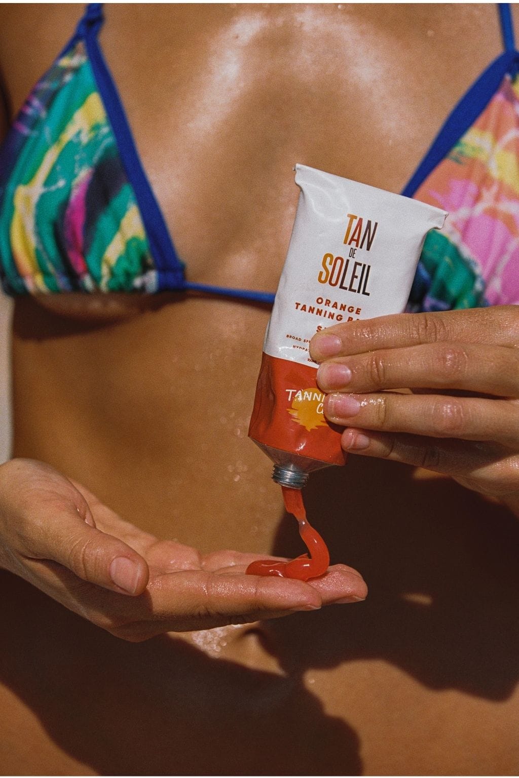 Person in colorful bikini holding Tan de Soleil orange tanning product tube and applying gel to hand.
