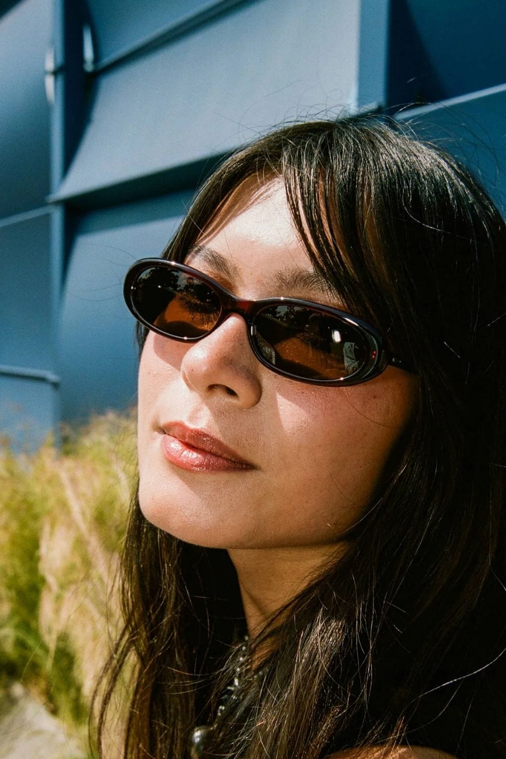 Black oval sunglasses with amber-tinted lenses worn by person with dark hair against blue wall.