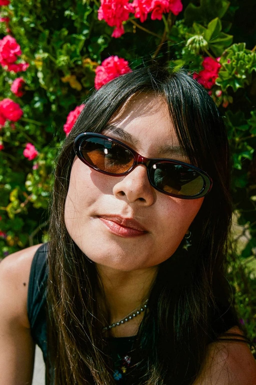 Person wearing oval tortoiseshell sunglasses with amber-tinted lenses against a backdrop of pink flowers and green foliage.