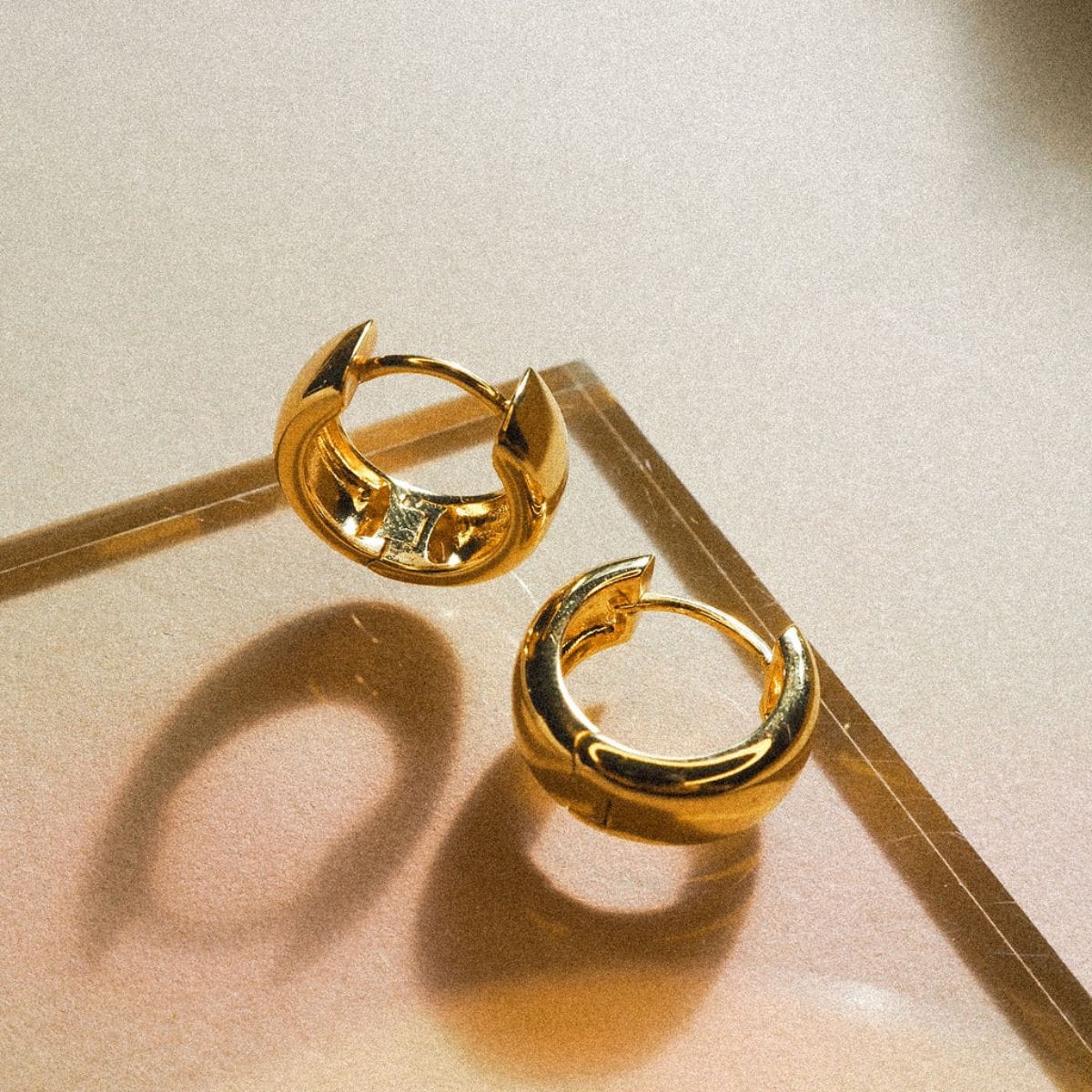 Gold-tone hoop earrings with pointed leaf-shaped ends, displayed on a neutral beige surface.