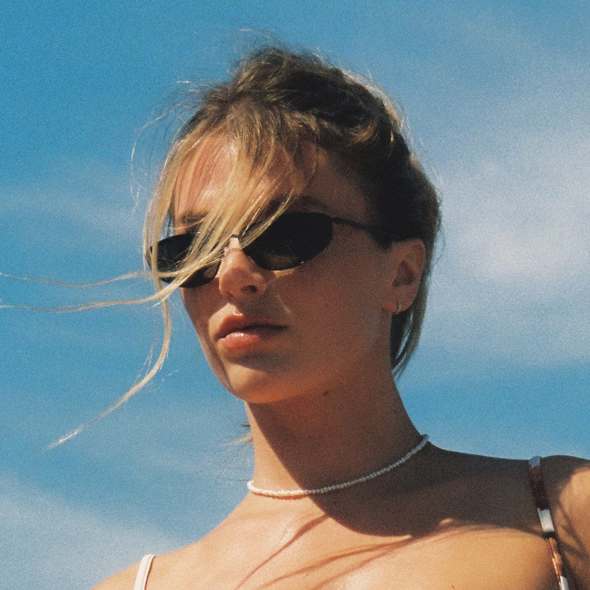 Dark oval sunglasses with thin metal frames worn by a person against a blue sky background.