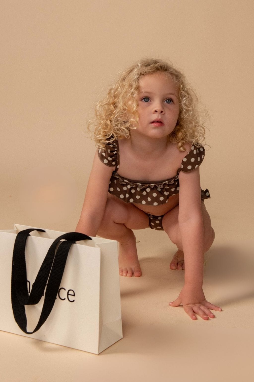 Toddler wearing brown and white polka dot bikini set with ruffled cap sleeves on neutral background.