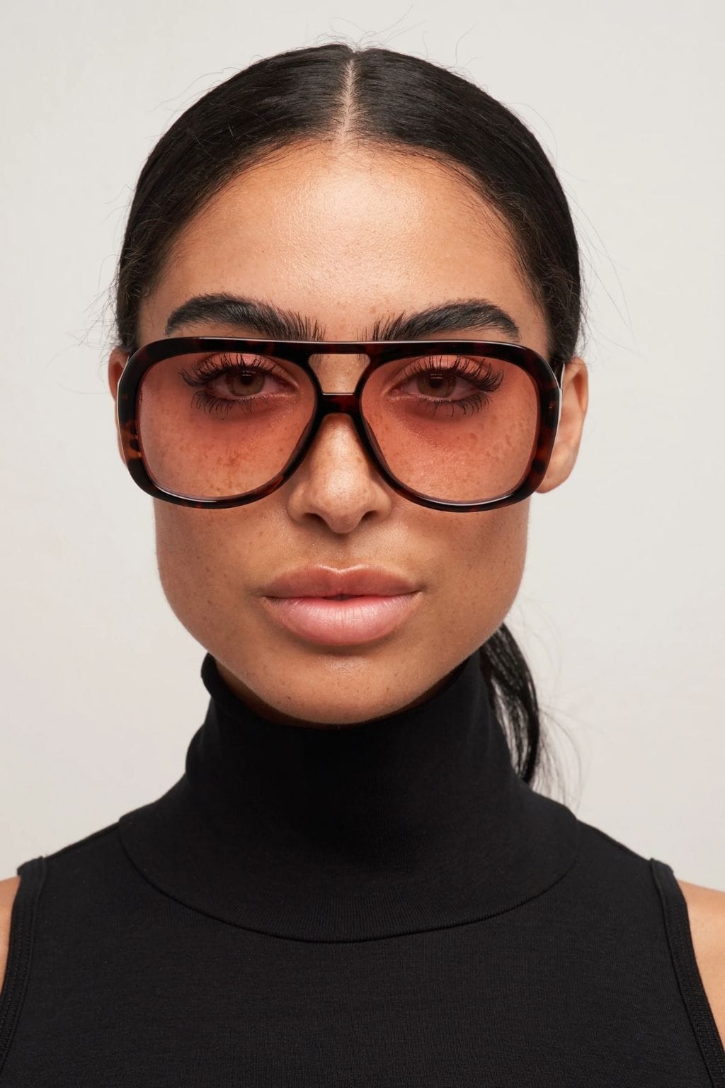 Oversized square sunglasses with tortoiseshell brown frames and pink-tinted lenses worn by a model in a black turtleneck.