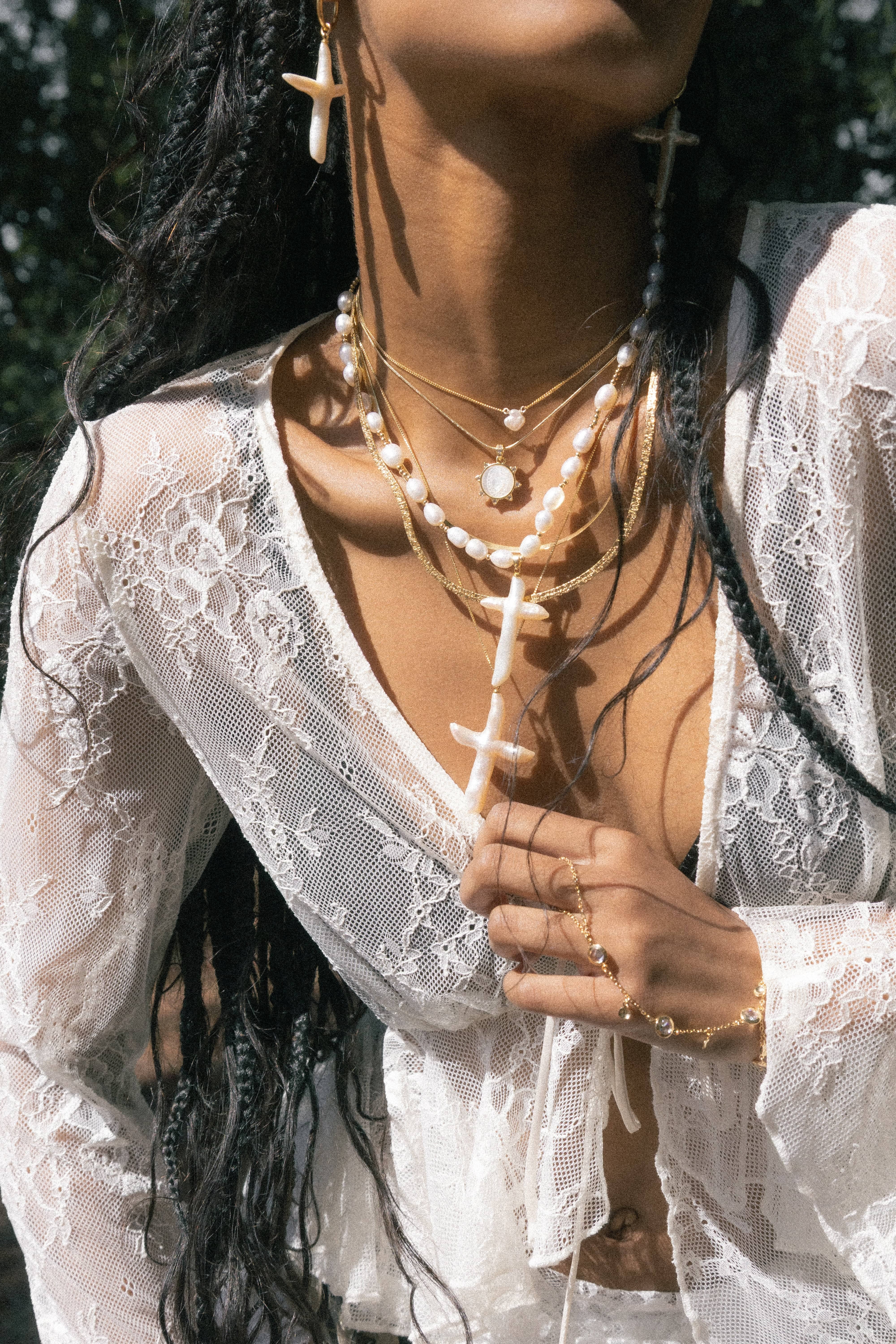 Gold chain necklace with white pearl beads and a white cross pendant, worn layered on a person's neck.