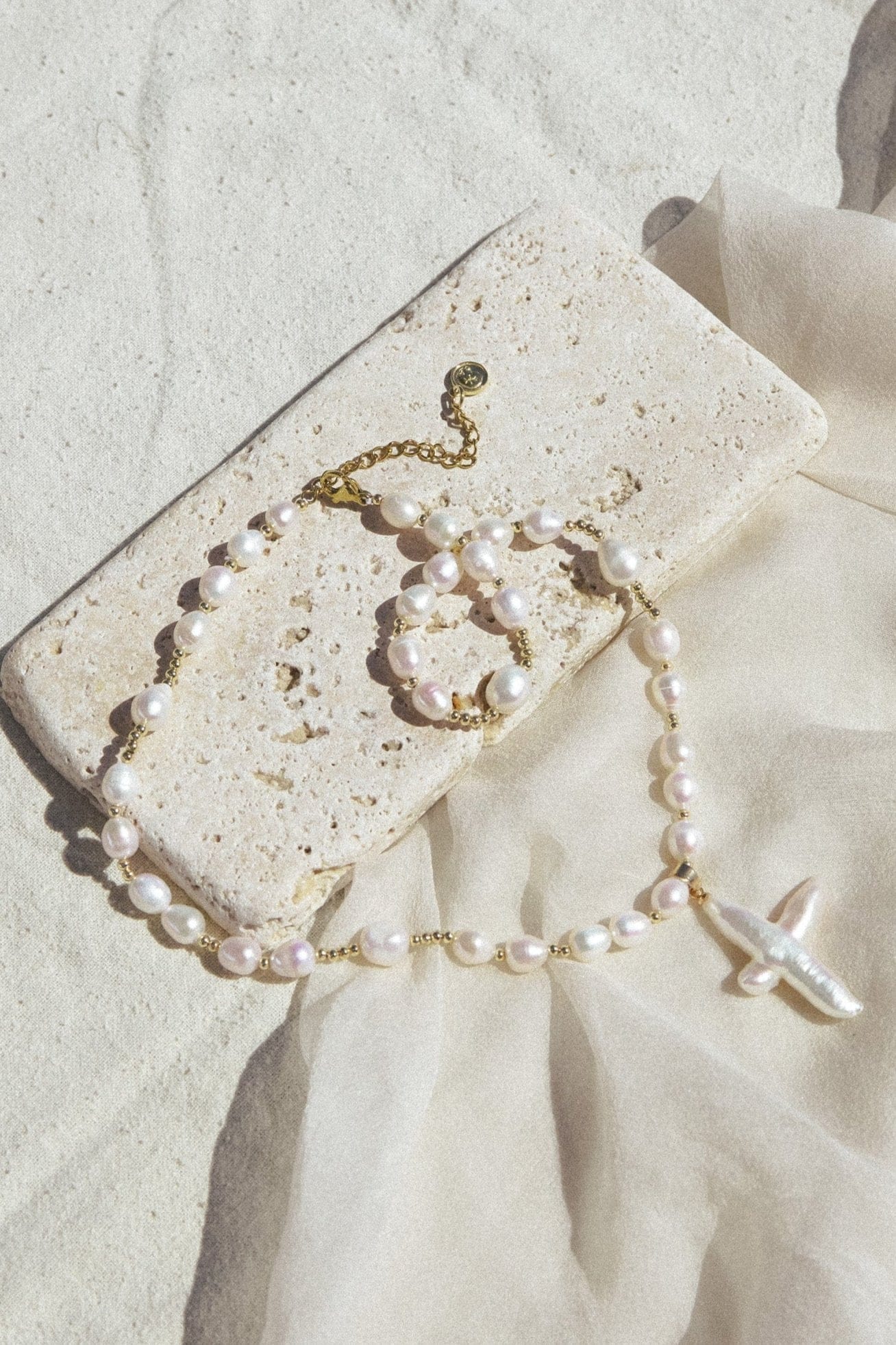 White pearl beaded necklace with gold-tone chain and white cross pendant displayed on textured stone.