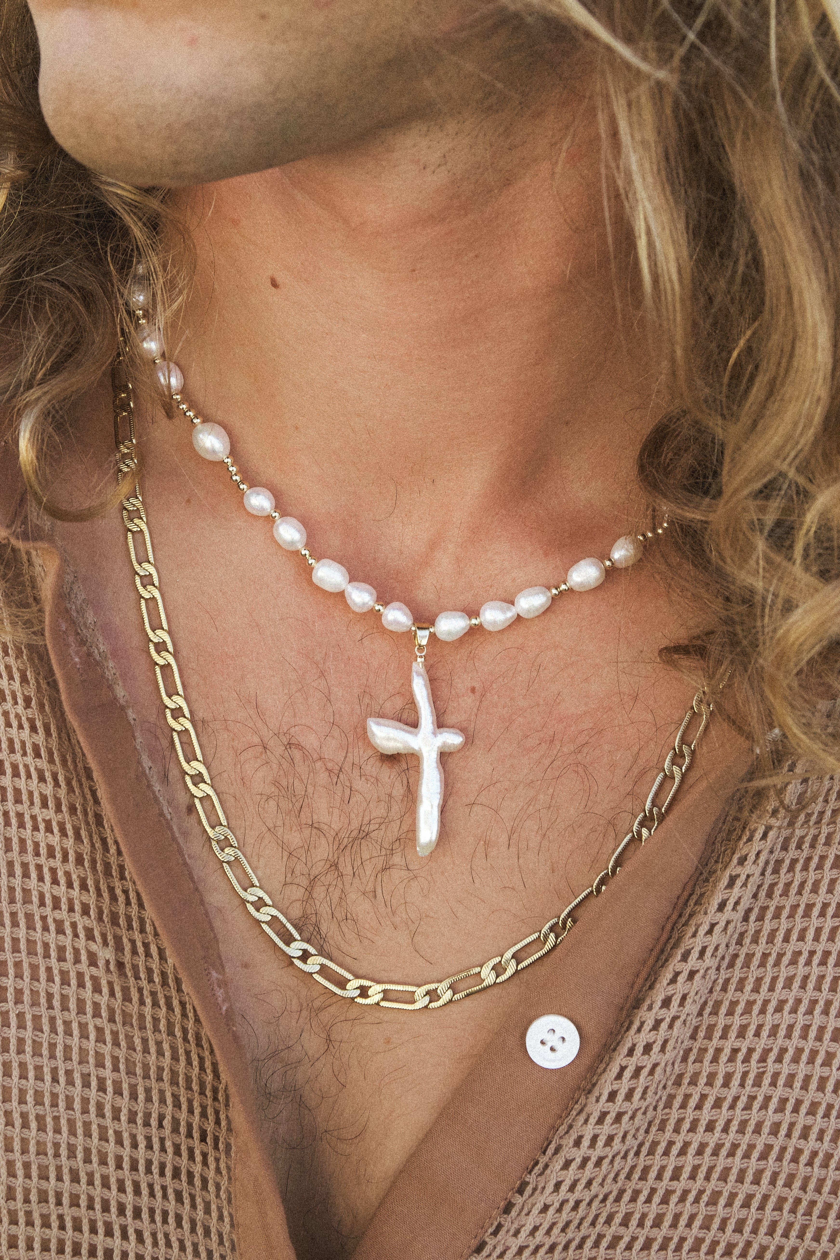 White pearl necklace with cross pendant worn layered with a gold chain on person's chest.
