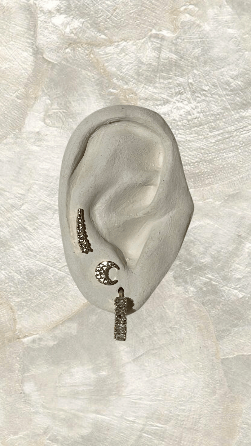 Silver-tone huggie earrings with cubic zirconia accents in crescent and bar designs on an ear model.