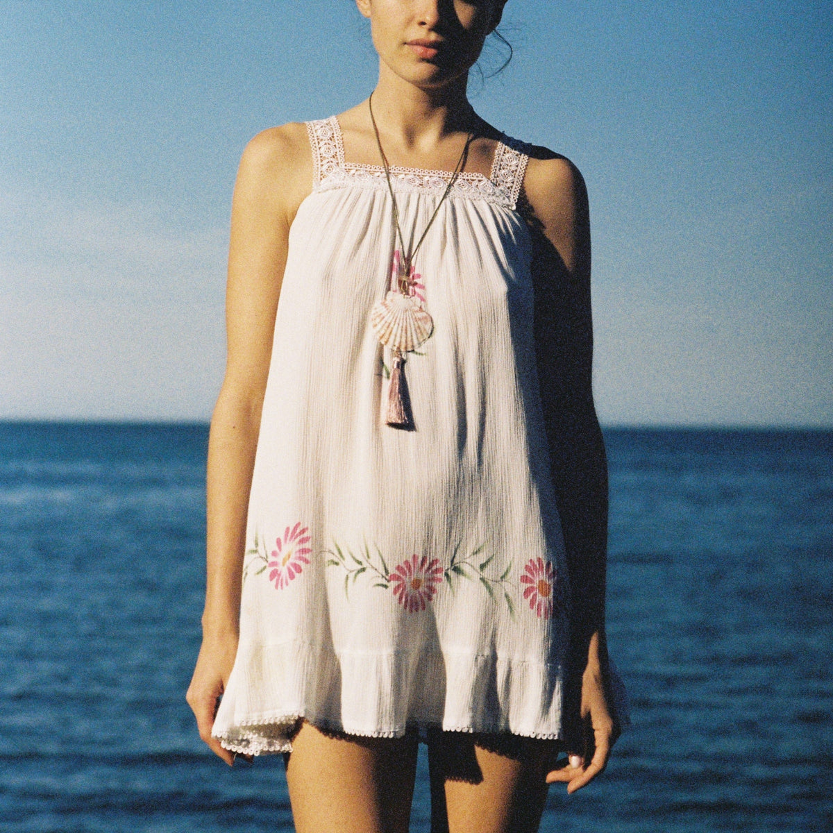 Long beaded necklace with shell and tassel pendants worn over white sleeveless dress.