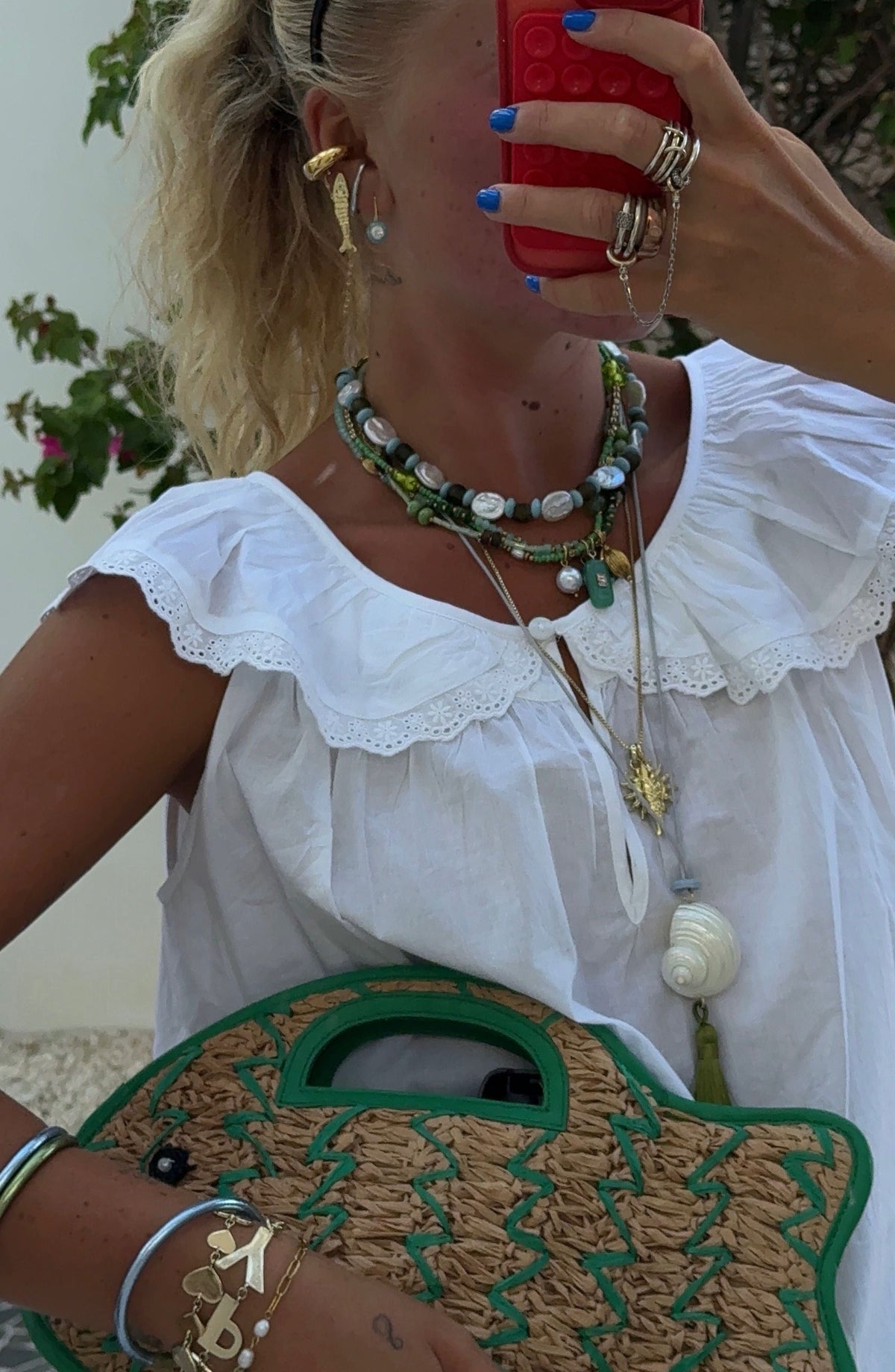 A person in a white eyelet-trimmed blouse wears layered green beaded and pearl necklaces, a shell pendant, and holds a woven green-trimmed bag.