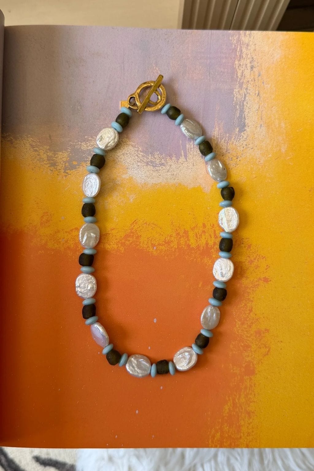 A beaded necklace with flat white pearls, dark olive beads, light blue spacers, and a gold toggle clasp lies on orange and yellow artwork.