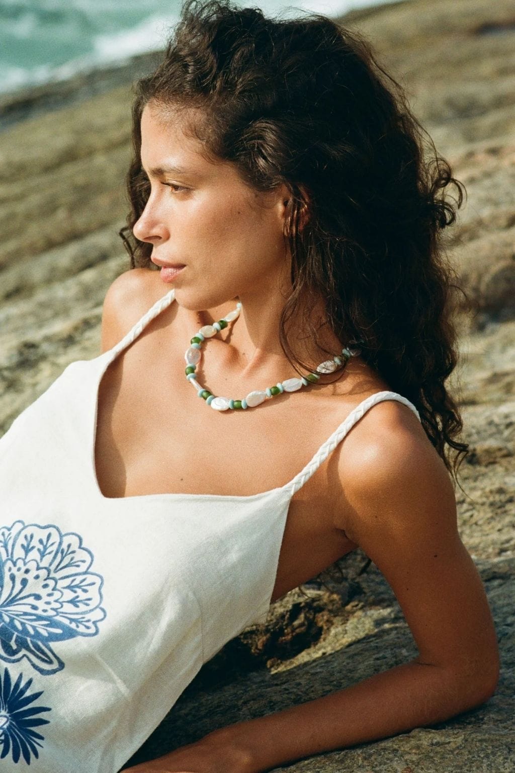 A woman with dark curly hair wears a beaded necklace of white shells and green and blue beads while reclining on rocks.