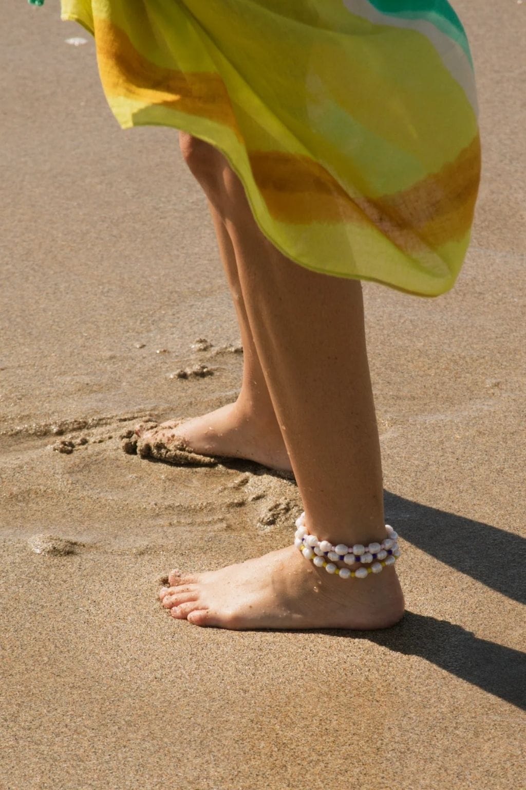 Person wearing multi-strand beaded anklet with white and yellow pearls on sandy beach.