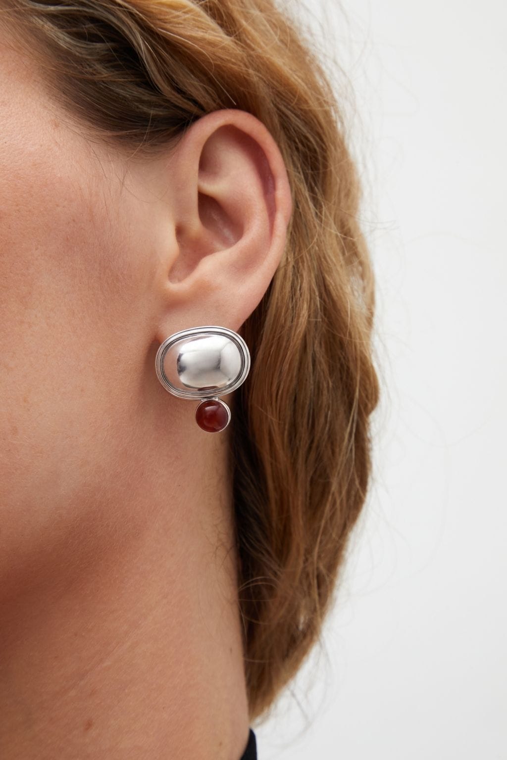 Silver earring with polished oval dome and small burgundy stone pendant worn on earlobe.