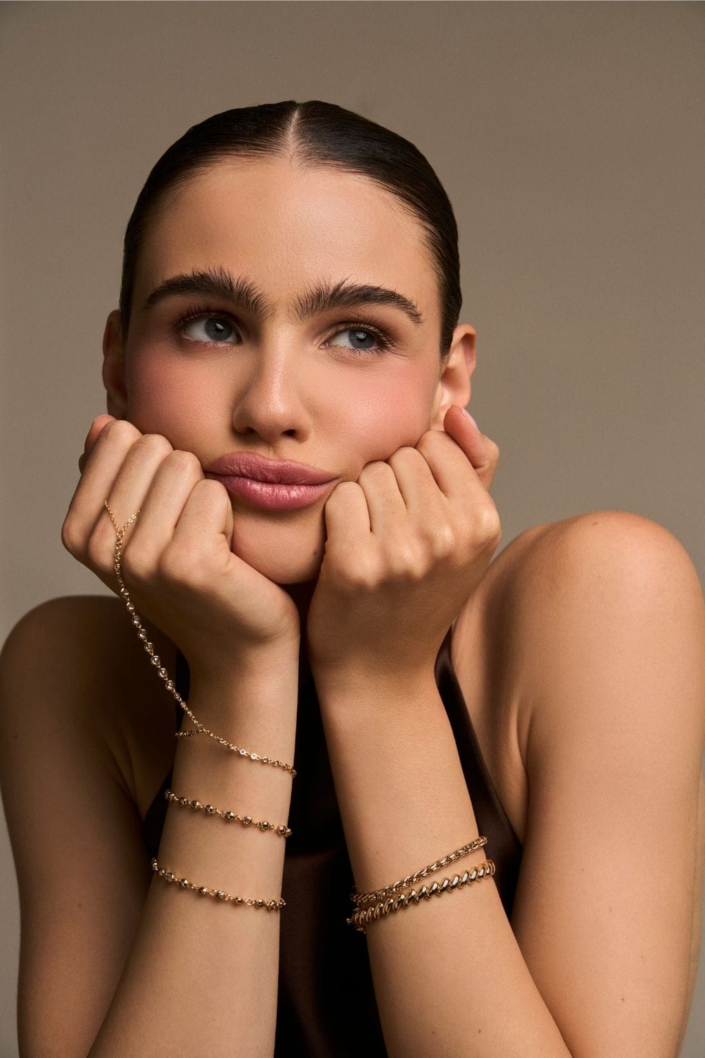 A woman rests her chin on both hands, wearing a gold beaded hand chain and several gold bracelets on her wrists.
