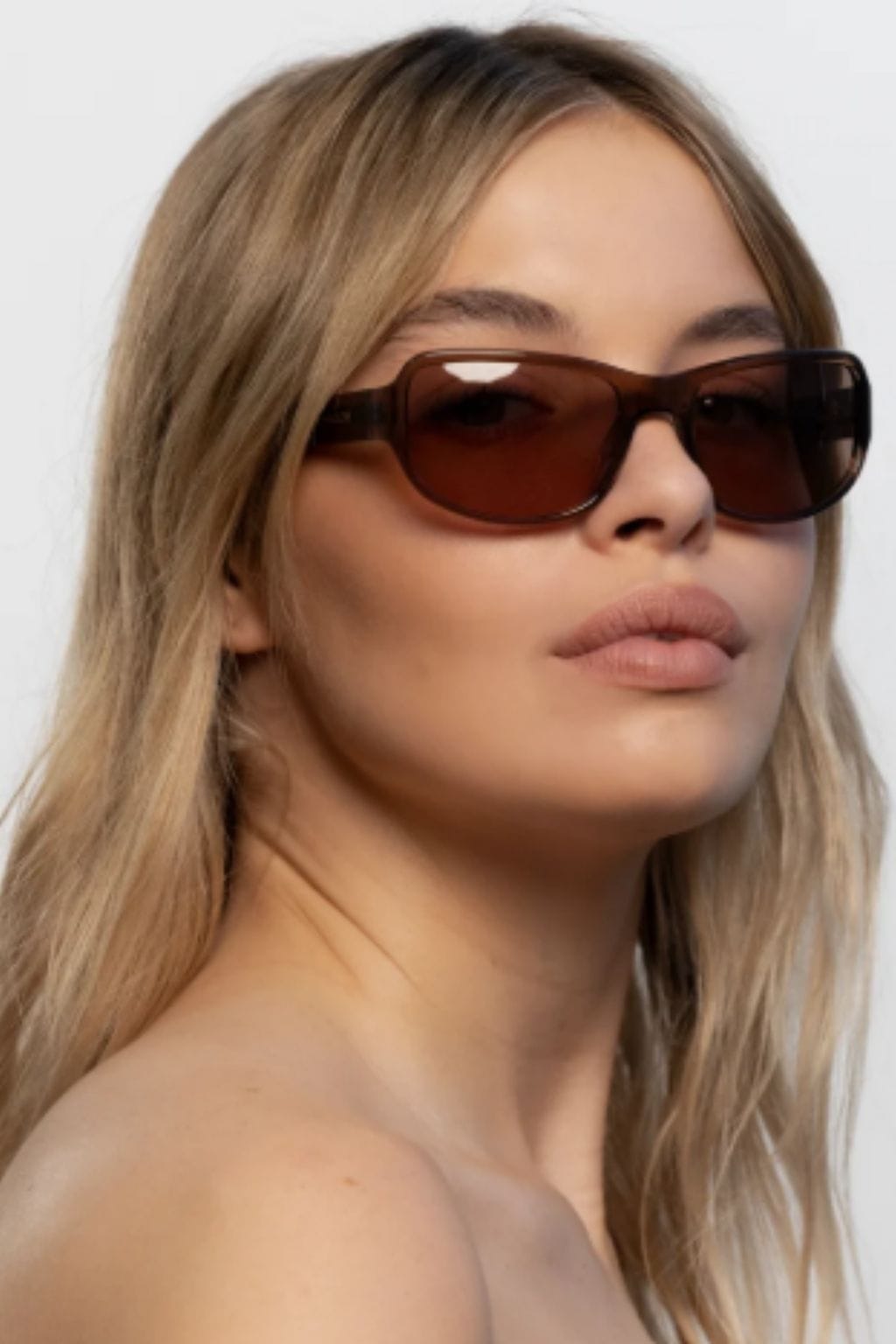 Woman wearing brown-tinted rectangular sunglasses with long blonde hair against white background.
