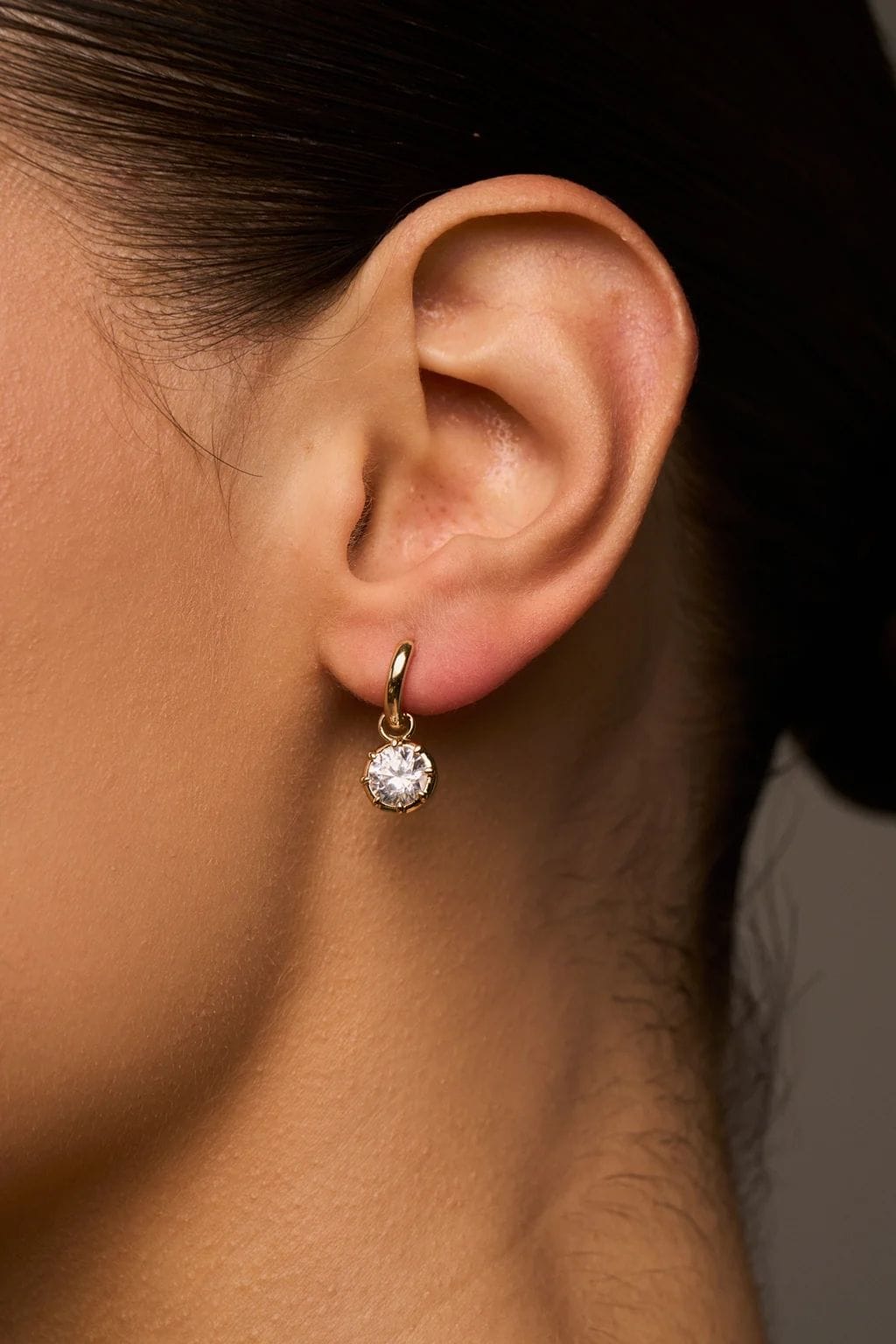 Gold-toned drop earring with round crystal stone worn on ear lobe.