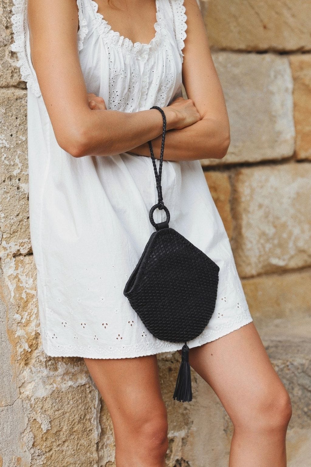 Black woven shoulder bag with tassel detail and cord strap worn by person in white dress.