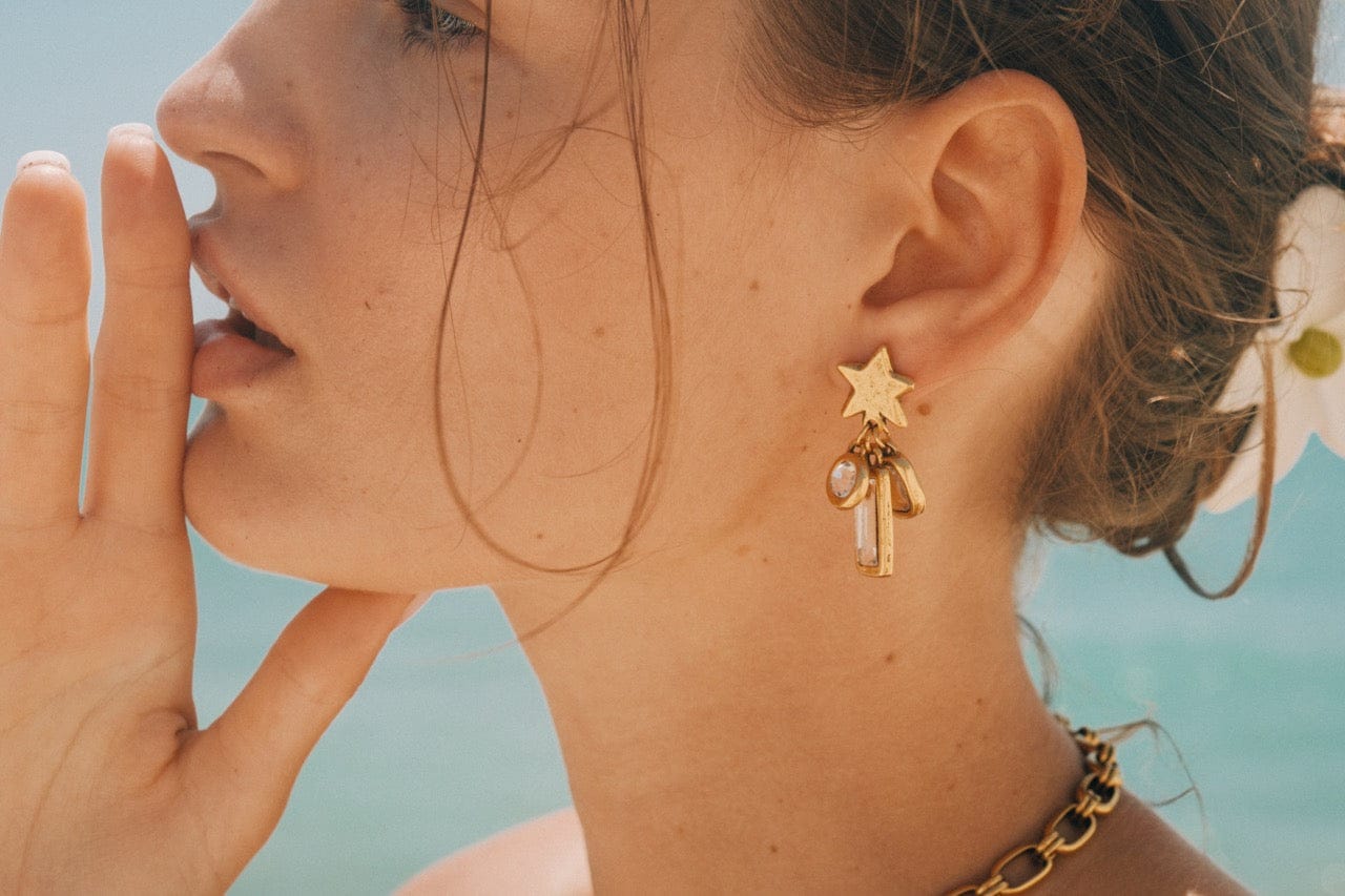 Gold-tone star earrings with dangling rectangular charm details, shown worn on model against blue sky background.