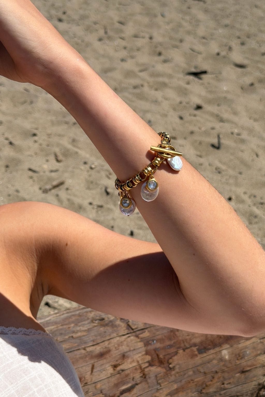 A gold chain bracelet with a toggle clasp and hanging pearl charms is worn on a bent forearm over sand.