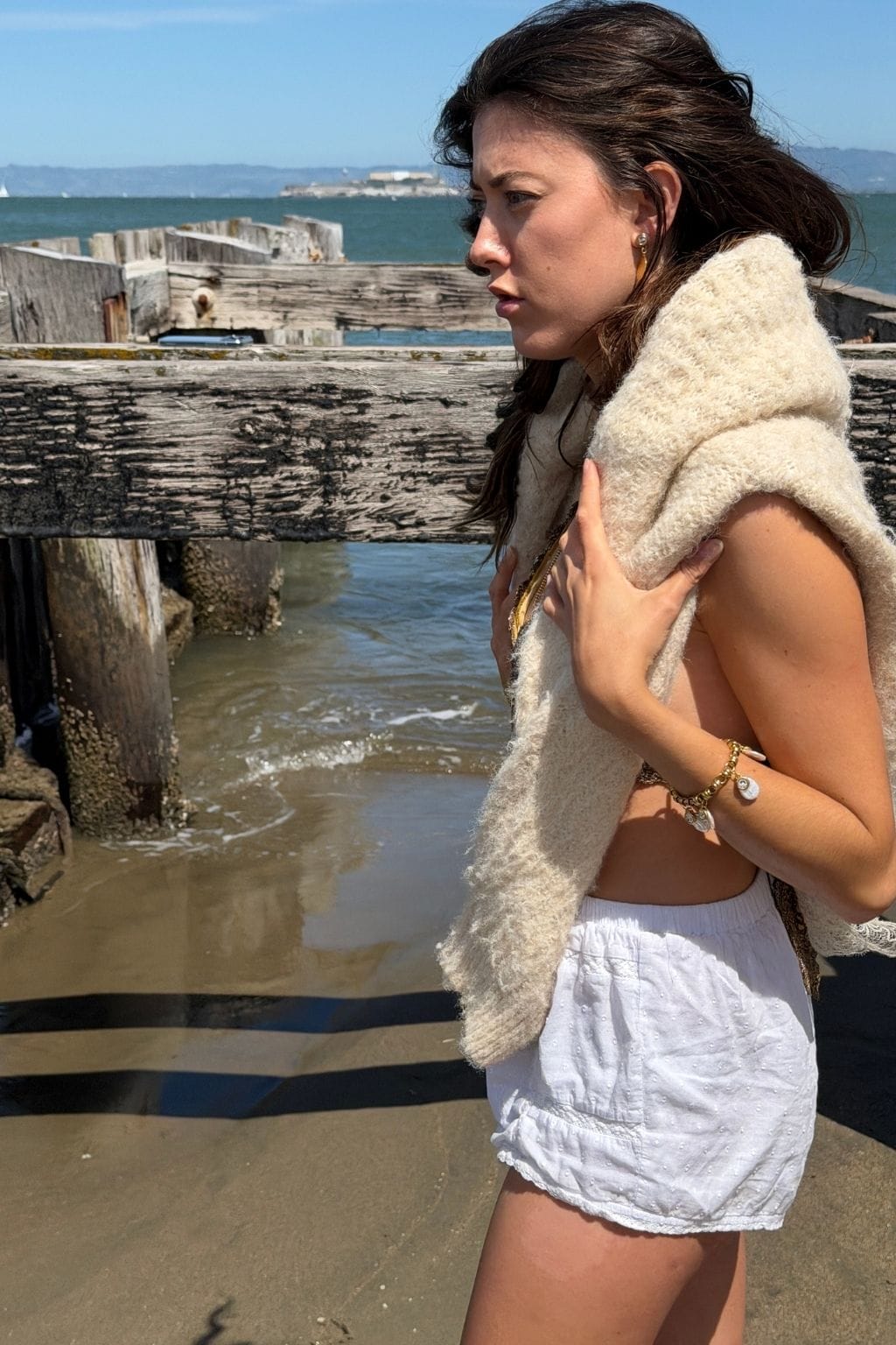 A woman on a sandy beach wears a gold chain bracelet with small charms, white shorts, and a cream knit shawl.