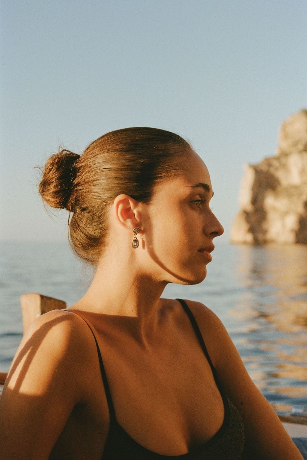 A woman with her hair in a bun wears small gold drop earrings in profile, with calm sea and rocky cliff behind.