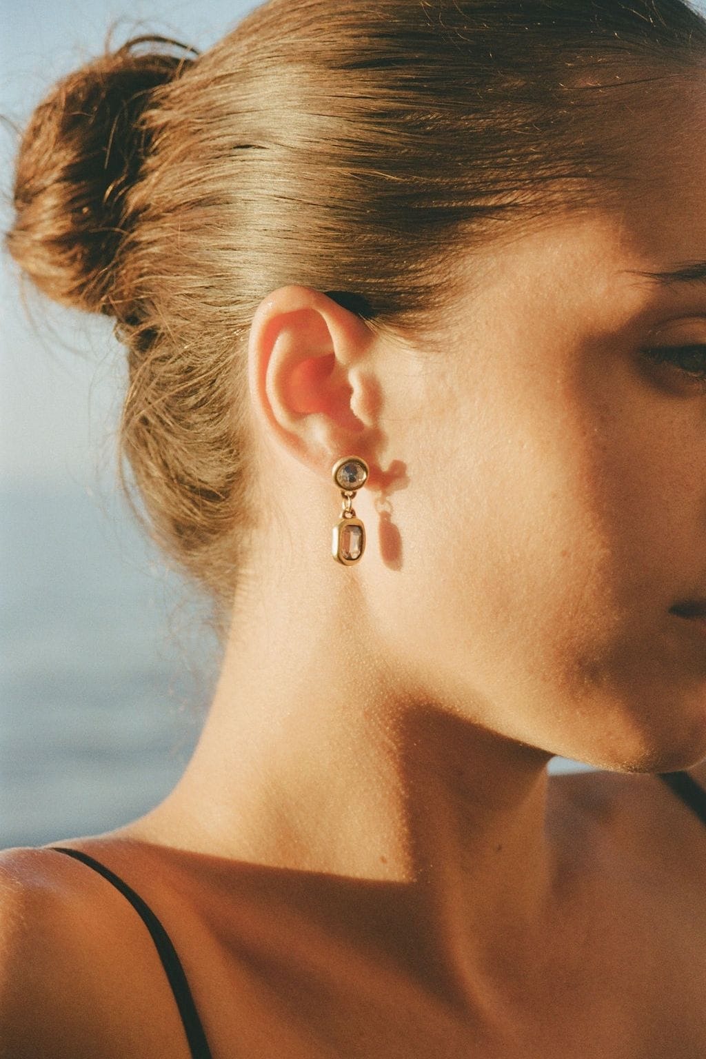 Side profile of a woman with her hair in a bun wearing a gold drop earring with two clear stones.
