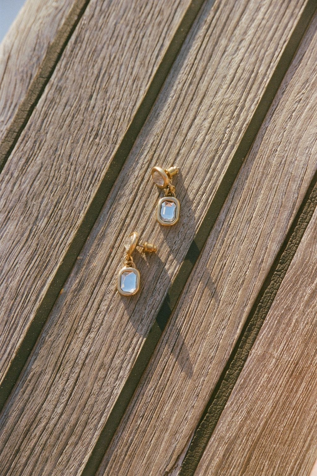 A pair of gold drop earrings with rectangular clear crystals rests on weathered wooden planks in warm sunlight.
