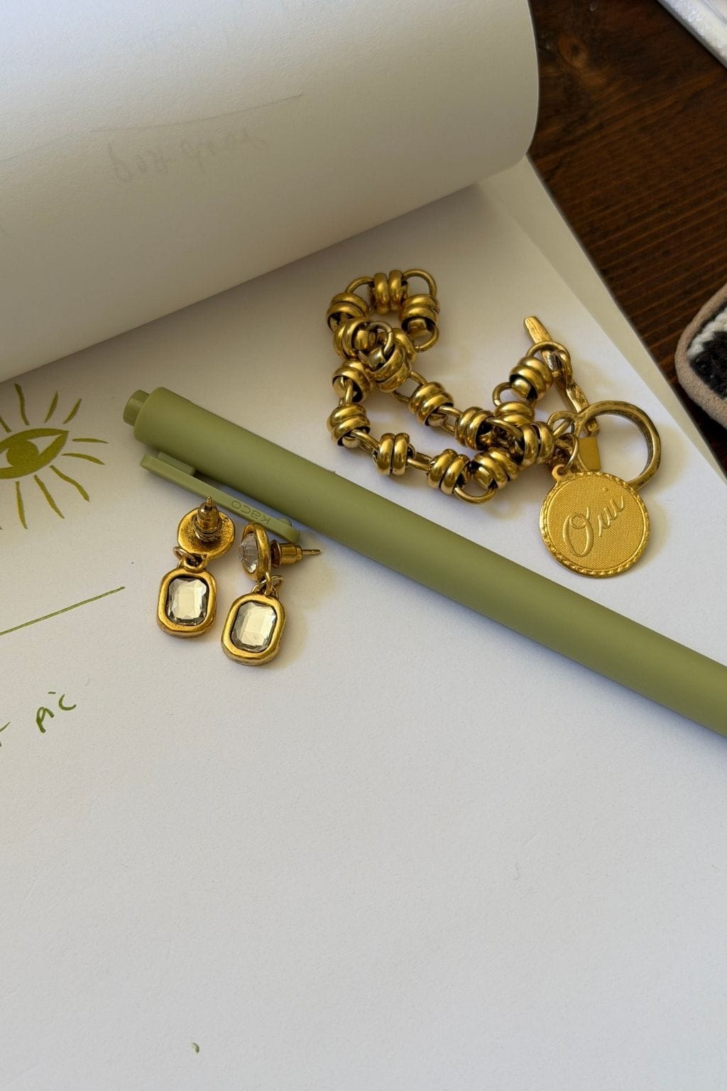 A pair of gold drop earrings with rectangular clear crystals rests on paper beside a green pen and gold chain bracelet.