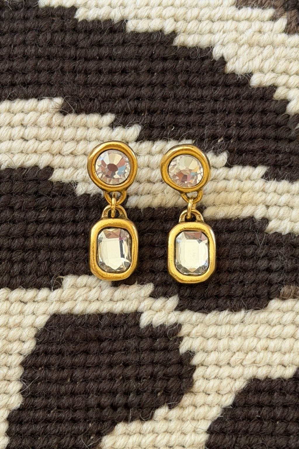 Gold drop earrings with a round clear crystal above a rectangular clear crystal rest on brown and cream woven fabric.