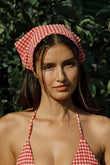 Red and white gingham scarf tied as headwrap on person with dark hair against green foliage background.