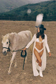 Woman wearing cream-colored open kimono with long sleeves over white swimsuit while leading a gray horse in a ranch setting.
