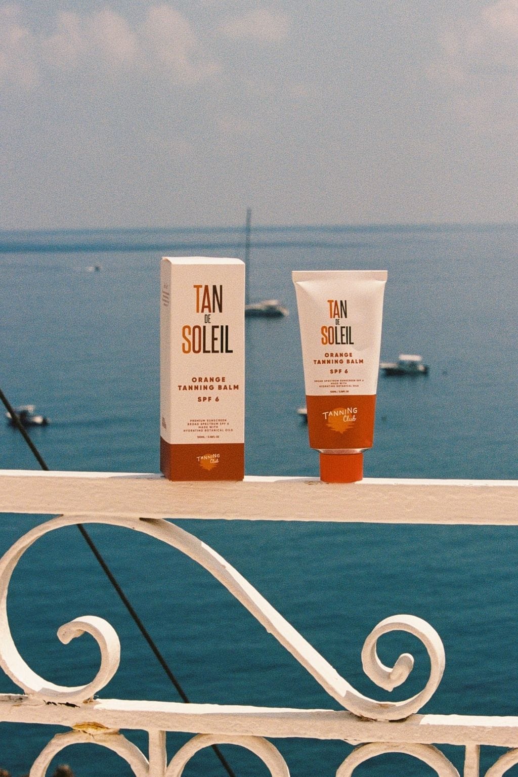 Tan de Soleil orange tanning balm SPF 6 in box and tube displayed on white railing with ocean backdrop.